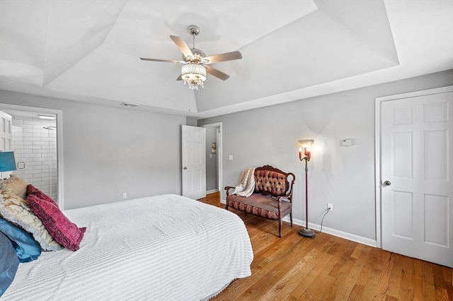 bedroom featuring a tray ceiling, light wood-style flooring, baseboards, ceiling fan, and ensuite bath