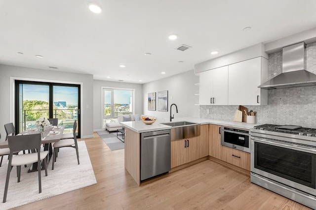 kitchen with light hardwood / wood-style floors, kitchen peninsula, wall chimney exhaust hood, appliances with stainless steel finishes, and light brown cabinets