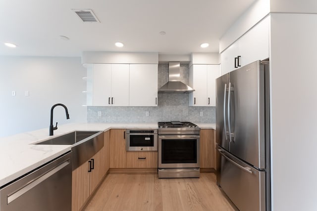 kitchen with appliances with stainless steel finishes, white cabinetry, backsplash, wall chimney exhaust hood, and light wood-type flooring