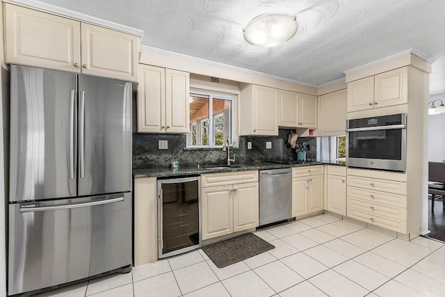 kitchen with cream cabinets, appliances with stainless steel finishes, a sink, beverage cooler, and tasteful backsplash