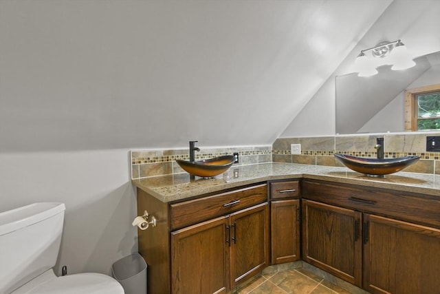 bathroom with lofted ceiling, toilet, double vanity, backsplash, and stone finish floors