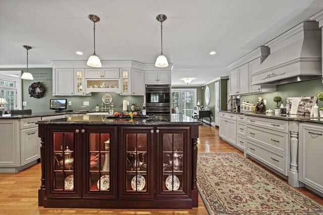 kitchen featuring a center island, custom range hood, hanging light fixtures, glass insert cabinets, and double oven