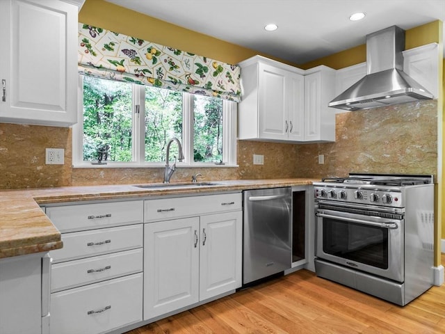 kitchen with wall chimney range hood, appliances with stainless steel finishes, white cabinetry, and a sink