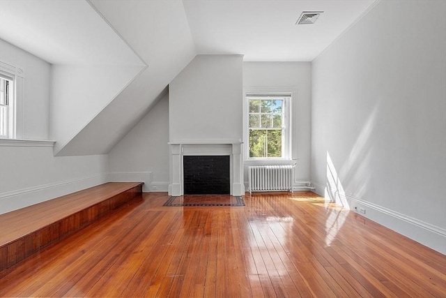 unfurnished living room featuring radiator, hardwood / wood-style floors, a fireplace with flush hearth, vaulted ceiling, and baseboards
