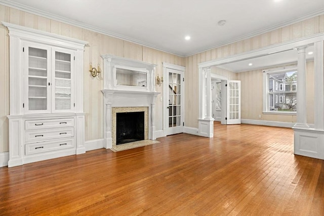 unfurnished living room with wood finished floors, ornamental molding, a fireplace with flush hearth, decorative columns, and baseboards