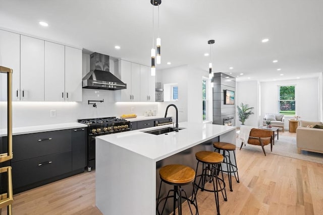 kitchen featuring wall chimney exhaust hood, sink, high end range, an island with sink, and pendant lighting