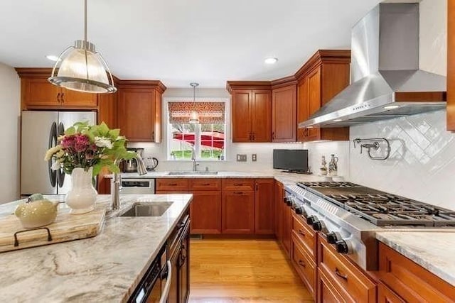 kitchen featuring wall chimney range hood, appliances with stainless steel finishes, hanging light fixtures, a sink, and light stone counters