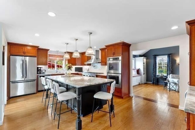 kitchen with appliances with stainless steel finishes, wall chimney range hood, a kitchen bar, a center island, and light wood-style flooring