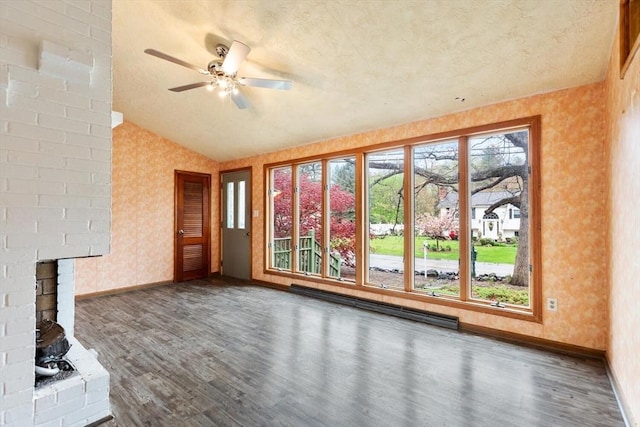 unfurnished living room featuring wood finished floors, baseboard heating, baseboards, a wood stove, and wallpapered walls