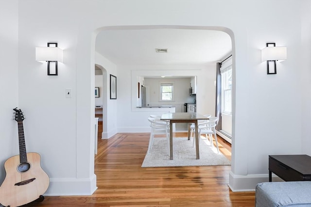 hall with arched walkways, light wood-type flooring, a baseboard radiator, and baseboards