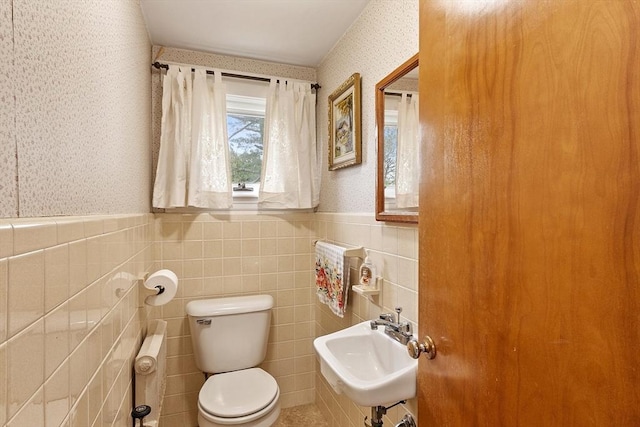 bathroom with wallpapered walls, toilet, a sink, wainscoting, and tile walls