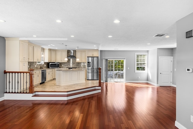 kitchen with cream cabinets, wall chimney range hood, light hardwood / wood-style flooring, stainless steel appliances, and hanging light fixtures