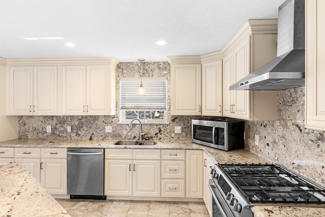 kitchen with wall chimney range hood, pendant lighting, cream cabinetry, sink, and appliances with stainless steel finishes