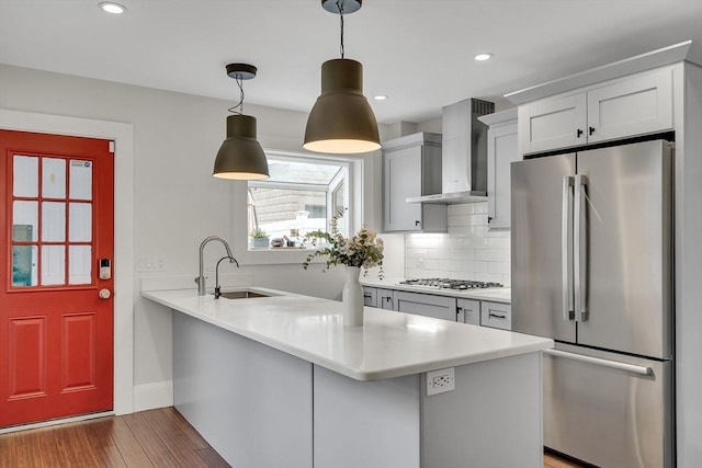 kitchen with a peninsula, a sink, wood finished floors, appliances with stainless steel finishes, and wall chimney range hood