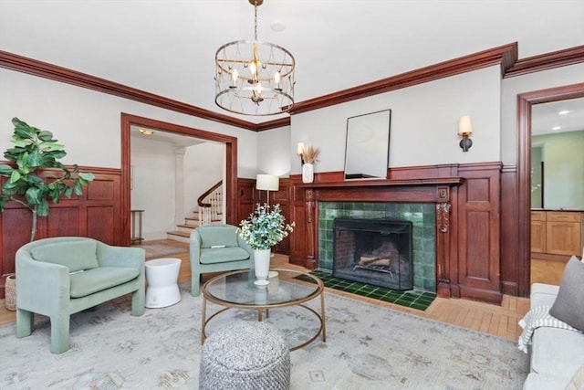 living room featuring a tile fireplace, stairs, ornamental molding, wainscoting, and an inviting chandelier