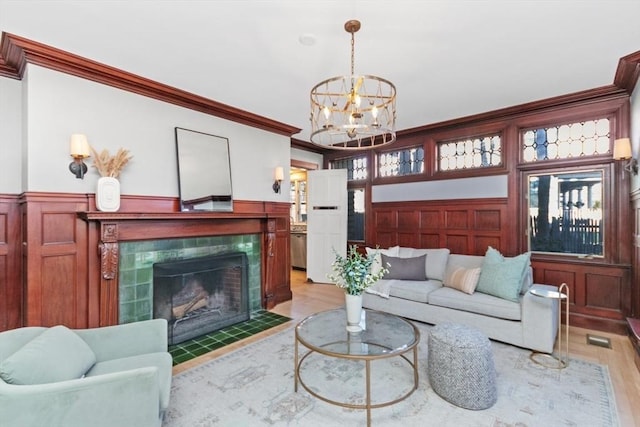 living area featuring light wood finished floors, a tiled fireplace, a wainscoted wall, crown molding, and a chandelier