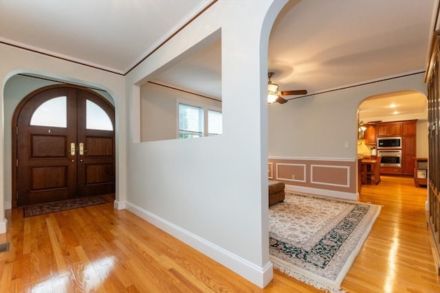 foyer with arched walkways, ornamental molding, a ceiling fan, light wood-style floors, and a wainscoted wall