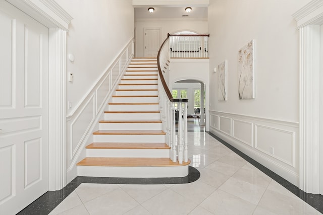 stairway with crown molding, arched walkways, a wainscoted wall, a high ceiling, and a decorative wall