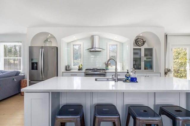 kitchen with decorative backsplash, white cabinets, stainless steel appliances, wall chimney exhaust hood, and a sink