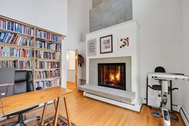 office featuring a glass covered fireplace, baseboard heating, wood finished floors, and a towering ceiling