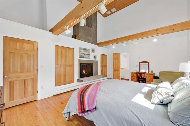 bedroom featuring track lighting, light wood finished floors, high vaulted ceiling, a glass covered fireplace, and beamed ceiling