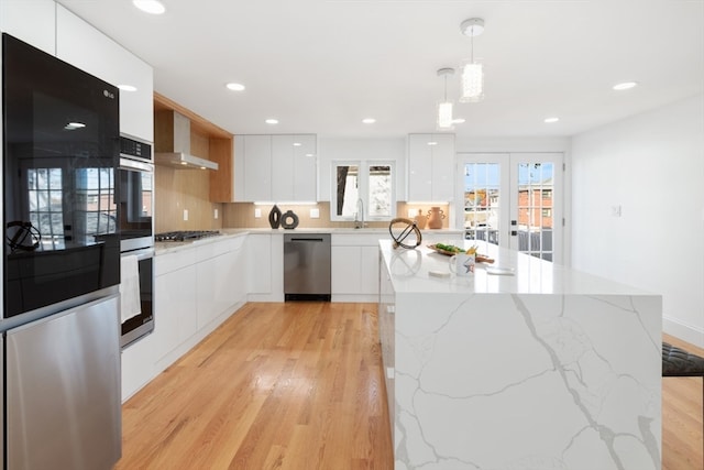 kitchen with appliances with stainless steel finishes, wall chimney range hood, light hardwood / wood-style flooring, white cabinets, and hanging light fixtures