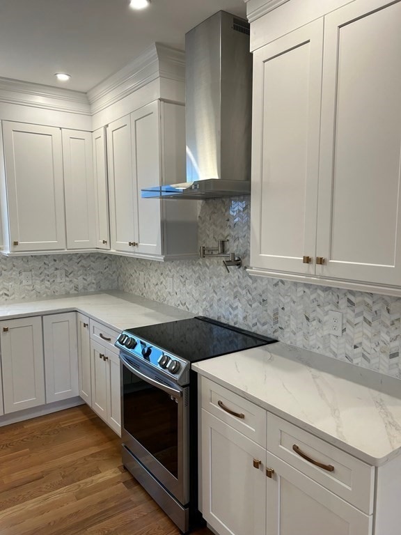 kitchen with wall chimney exhaust hood, hardwood / wood-style floors, stainless steel electric range oven, backsplash, and white cabinetry