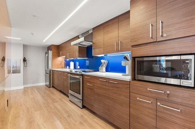 kitchen featuring light wood finished floors, wall chimney exhaust hood, appliances with stainless steel finishes, and brown cabinets