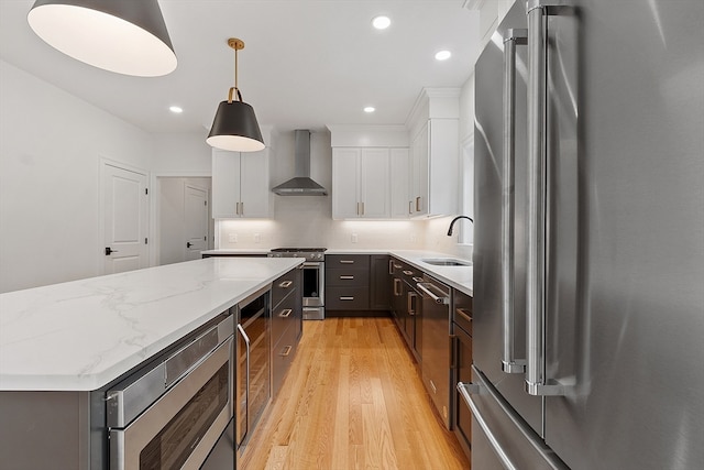 kitchen featuring appliances with stainless steel finishes, hanging light fixtures, light hardwood / wood-style floors, white cabinets, and wall chimney range hood