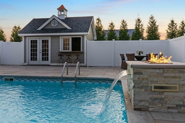 pool at dusk with pool water feature, an outdoor structure, an outdoor bar, a fire pit, and a patio