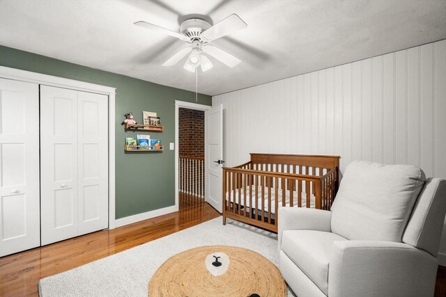 bedroom with wood finished floors, a nursery area, a closet, baseboards, and a ceiling fan