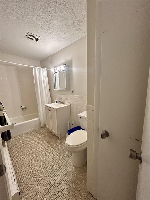 full bathroom with visible vents, tile walls, shower / bath combo with shower curtain, toilet, and a textured ceiling