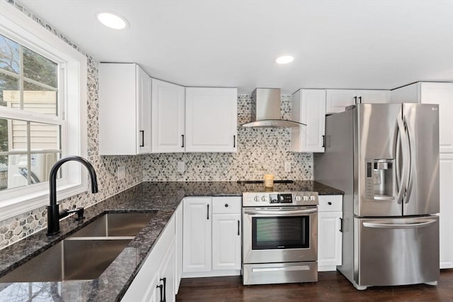 kitchen featuring appliances with stainless steel finishes, white cabinets, wall chimney range hood, dark stone countertops, and sink