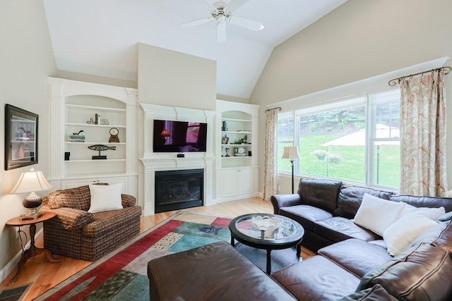 living area with wood finished floors, a ceiling fan, a glass covered fireplace, built in shelves, and high vaulted ceiling