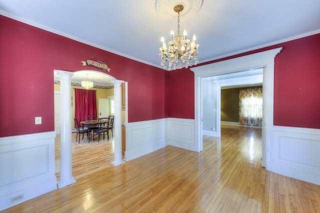 unfurnished dining area featuring crown molding, an inviting chandelier, a wainscoted wall, light wood-type flooring, and ornate columns