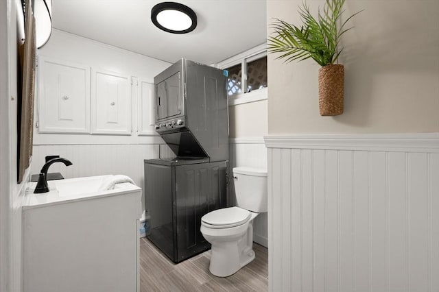 half bathroom with stacked washer and clothes dryer, toilet, wainscoting, vanity, and wood finished floors