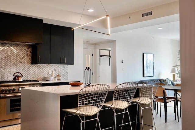 kitchen featuring dark cabinetry, high end range, a breakfast bar area, ventilation hood, and tasteful backsplash