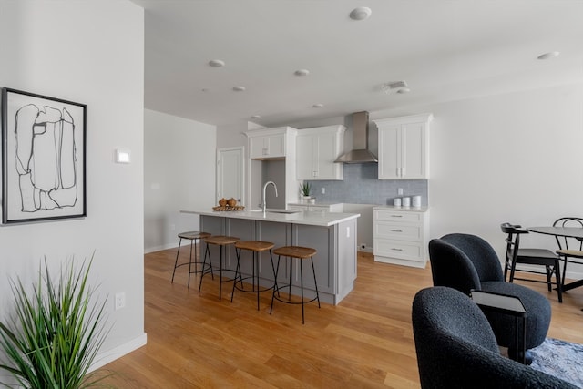 kitchen featuring a kitchen breakfast bar, wall chimney exhaust hood, light hardwood / wood-style flooring, a kitchen island with sink, and white cabinetry