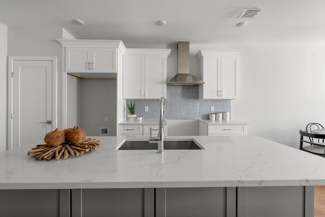 kitchen with tasteful backsplash, a kitchen island with sink, sink, light stone counters, and wall chimney range hood