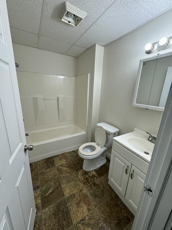 full bath featuring toilet, stone finish flooring, vanity, a paneled ceiling, and washtub / shower combination