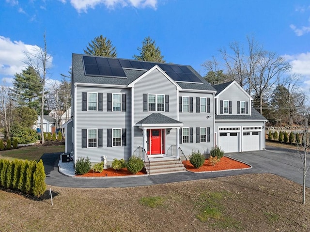 colonial house with an attached garage, roof mounted solar panels, driveway, and roof with shingles