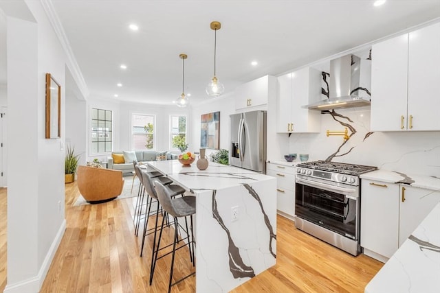 kitchen with wall chimney range hood, stainless steel appliances, crown molding, pendant lighting, and light wood-style flooring