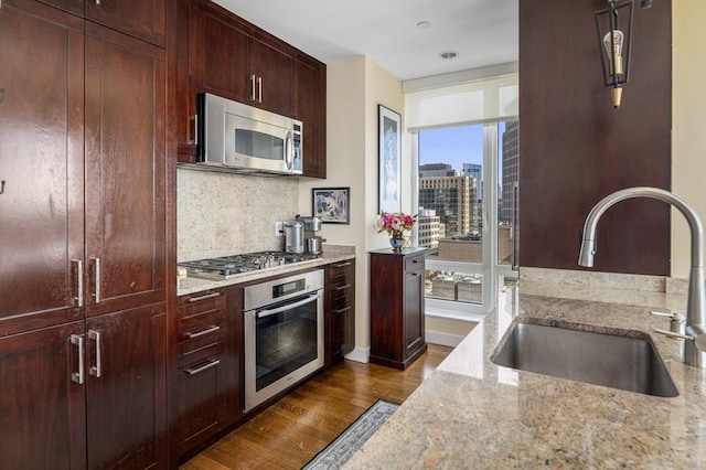 kitchen with light stone counters, stainless steel appliances, a sink, backsplash, and dark wood-style floors