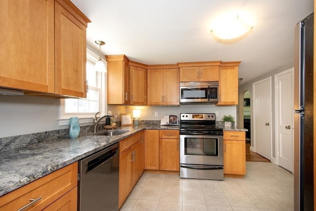 kitchen with appliances with stainless steel finishes, brown cabinets, a sink, light tile patterned floors, and dark stone counters