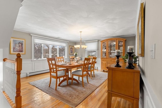 dining room with a notable chandelier, ornamental molding, baseboard heating, wainscoting, and light wood finished floors