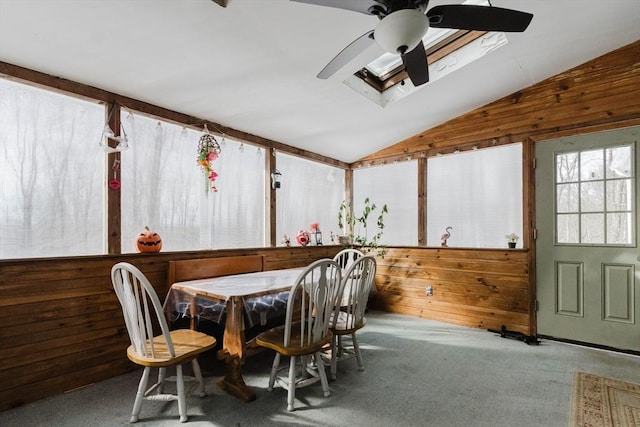 dining space featuring vaulted ceiling with skylight, wood walls, carpet, and a ceiling fan