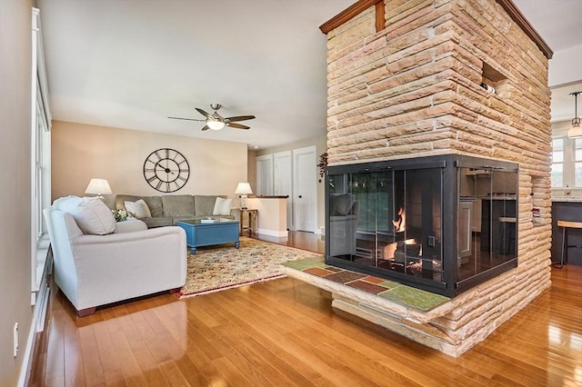 living room with wood-type flooring, a ceiling fan, baseboards, and a stone fireplace
