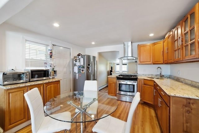 kitchen featuring wall chimney range hood, baseboard heating, appliances with stainless steel finishes, brown cabinetry, and glass insert cabinets