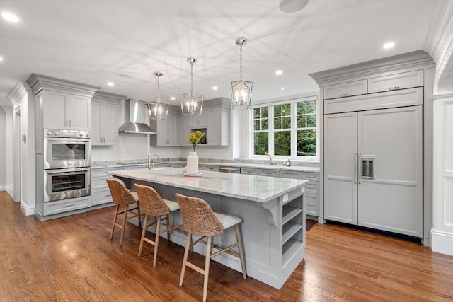 kitchen with light stone counters, gray cabinetry, appliances with stainless steel finishes, wall chimney exhaust hood, and an island with sink