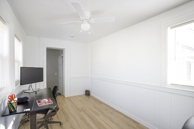 office with light wood finished floors, healthy amount of natural light, a wainscoted wall, a ceiling fan, and a decorative wall